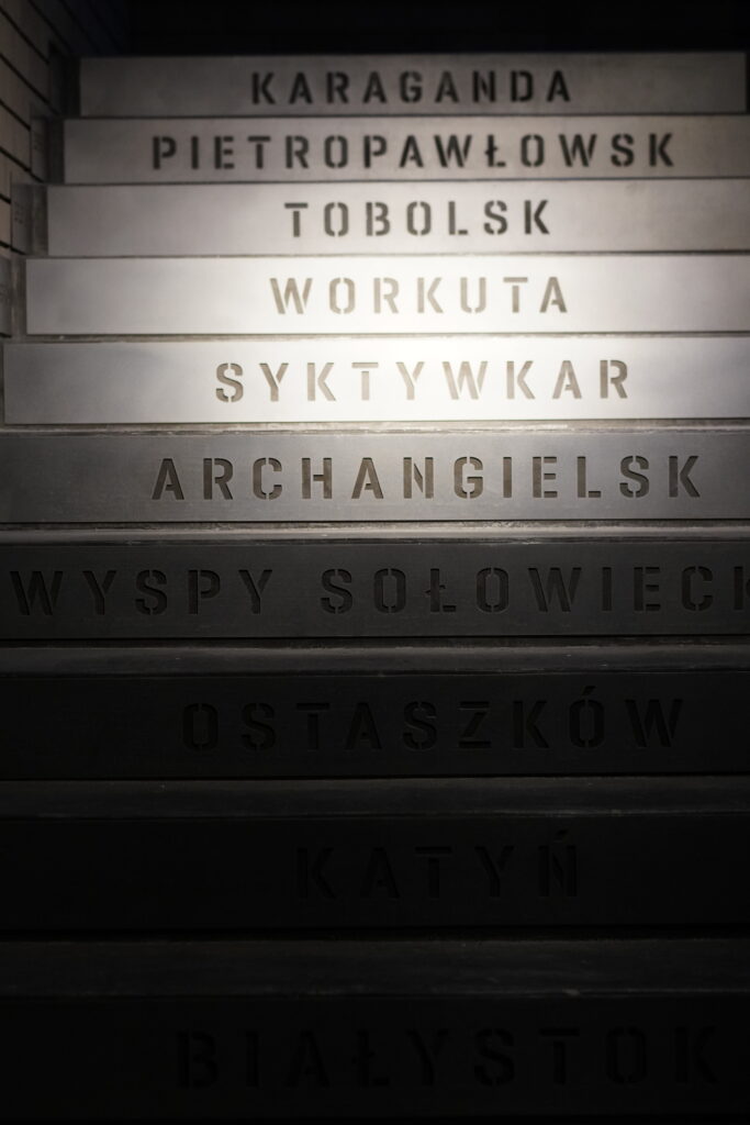 Zdjęcie przedstawia nazwy miast umieszczone na schodach prowadzących na białą salę wystawy stałej Muzeum.
