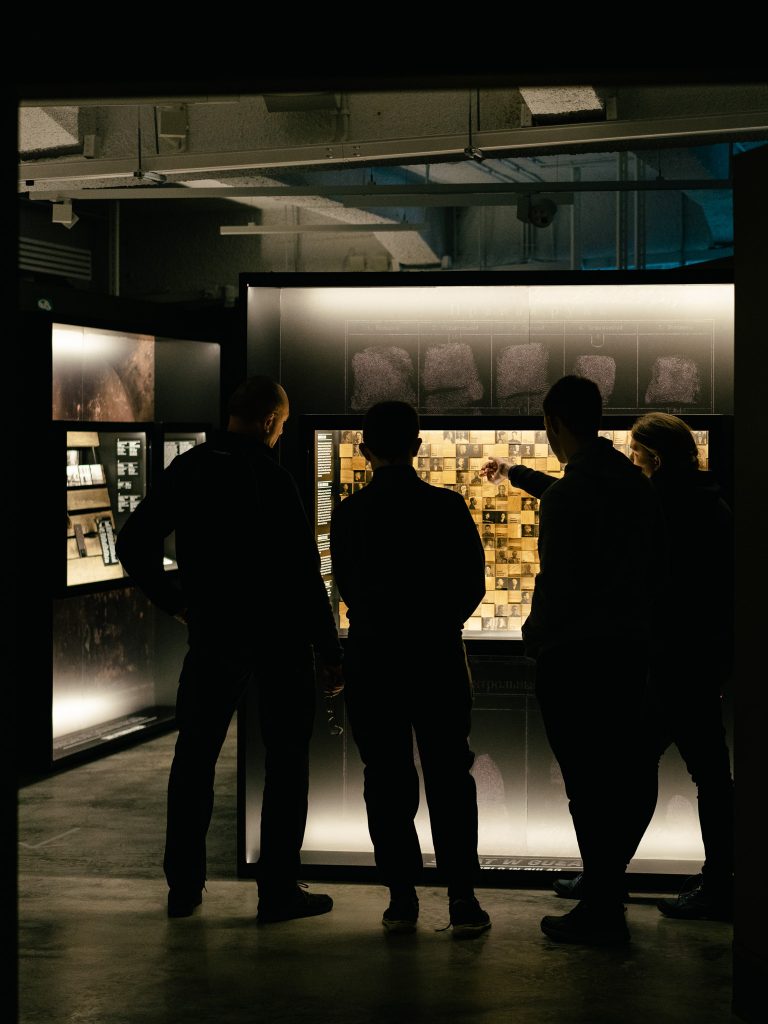 Cztery osoby stojące przy podświetlonej gablocie podczas Pierwszego Syberyjskiego Salonu Ambientu. Oglądają przedstawione fotografie.