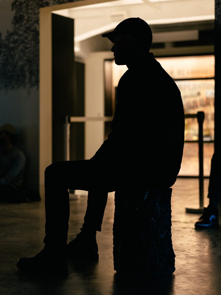 Mężczyzna w czapce siedzący na stołku podczas Pierwszego Syberyjskiego Salonu Ambientu. Patrzy przed siebie.