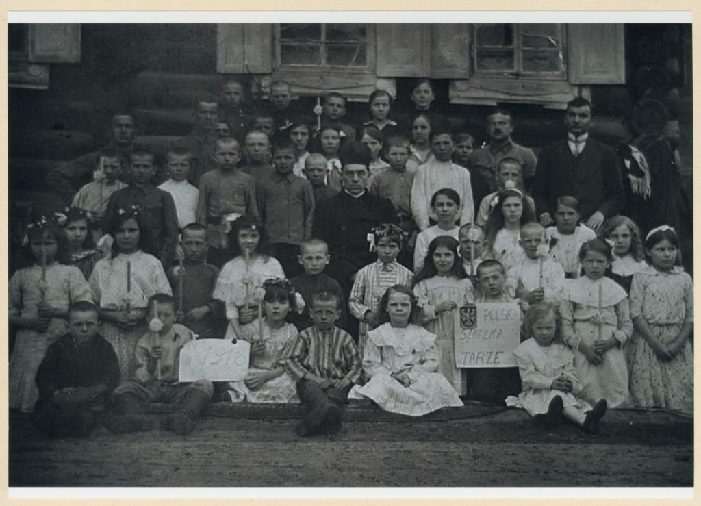 Czarno-białe zdjęcie przedstawiające grupę stojących, siedzących lub klęczących dzieci. W środku grupy siedzi ksiądz. Część dzieci trzyma w rękach długie świece oraz kartki z napisami: "Polska szkółka w Tarze" oraz "1918".