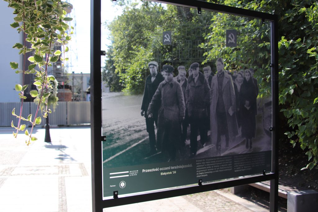 Jeden z pięciu elementów wystawy plenerowej "Przeszłość oczami teraźniejszości. Białystok' 39". W ramie przezroczysta tafla z nadrukowanym archiwalnym, czarno-białym zdjęciem. Przez niezadrukowany fragment tafli przebija widok ulicy. Współczesny widok uzupełnia archiwalną fotografię.