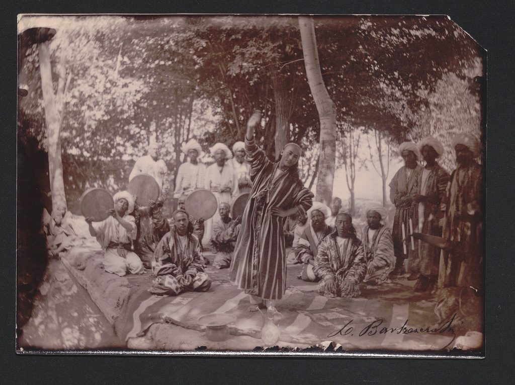 Zdjęcie w odcieniach sepii, wykonane przez Leona Barszczewskiego, przedstawia przedstawiające tancerza w pasiastej szacie, tańczącego przy wtórze bębnów, na których grają klęczący wokół mężczyźni.