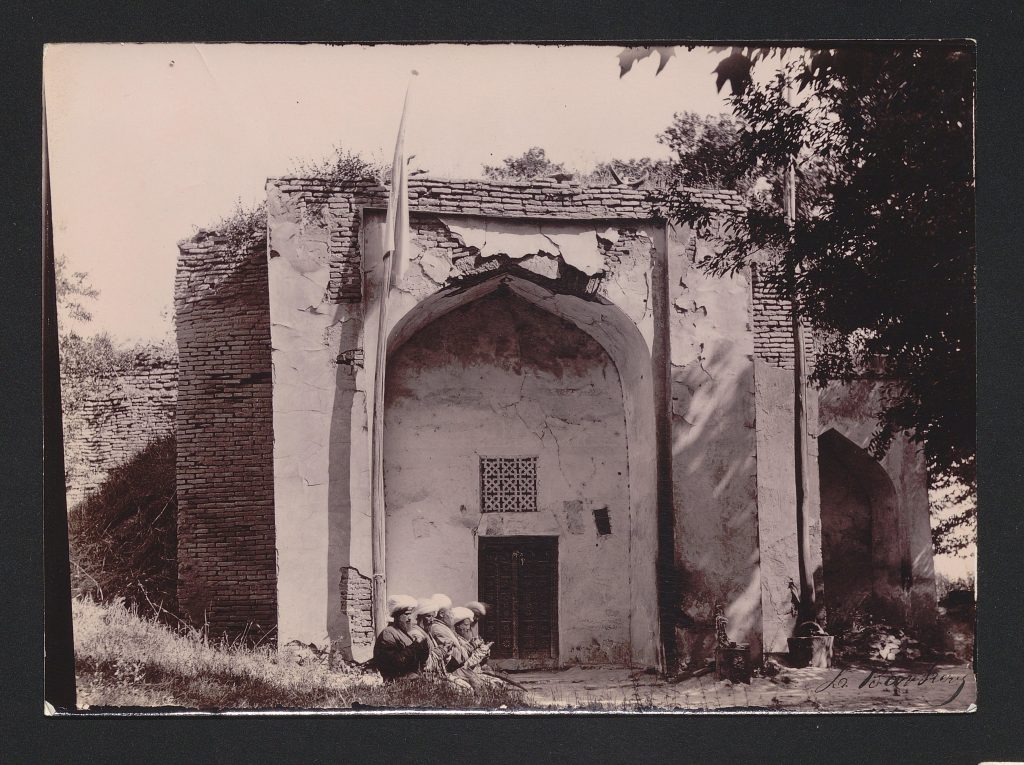 Zdjęcie w kolorze sepii, wykonane przez Leona Barszczewskiego, przedstawia częściowo zrujnowany meczet z wejściem w głębokiej niszy. Na pierwszym planie grupa modlących się mężczyzn w białych zawojach na głowach. Fotografia wykonana przez Leona Barszczewskiego w latach 70.-90. XIX wieku