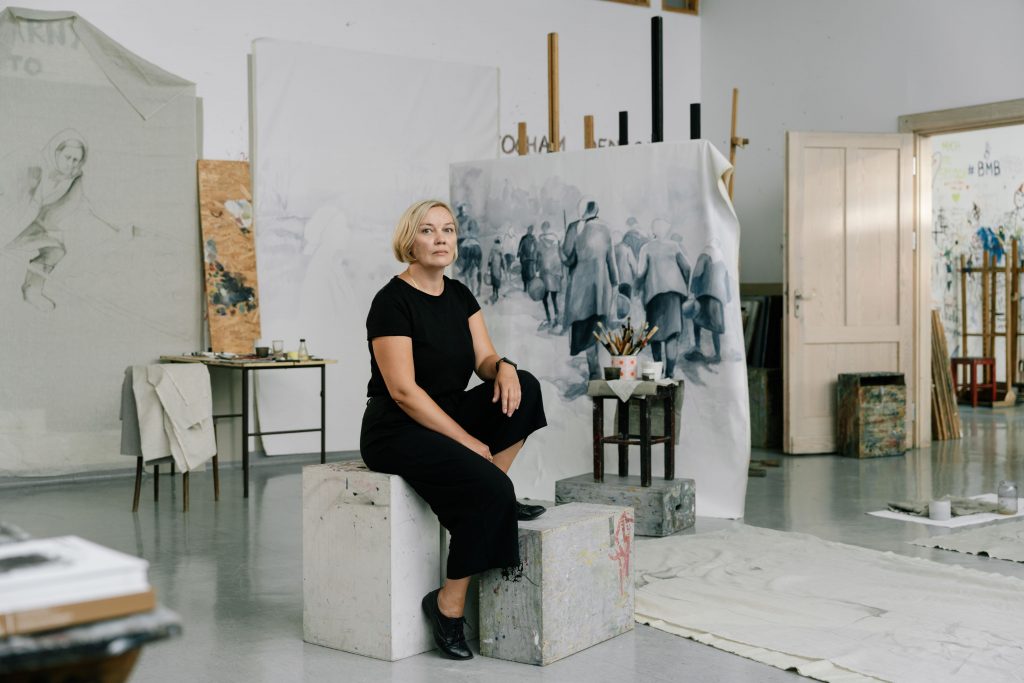 Edyta Urwanowicz siedzi w swojej pracowni malarskiej. Za artystką płótno z obrazem w szarej tonacji.