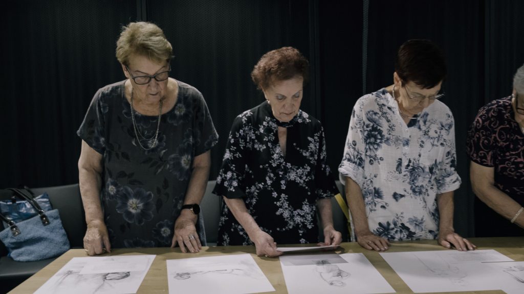 Trzy starsze kobiety patrzą na czarno-białe rysunki rozłożone przed nimi na stole.