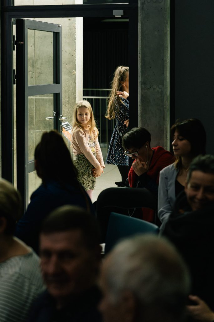 Publiczność na spotkaniu z podróżnikiem Krzysztofem Suchowierskim. W drzwiach, w plamie ciepłego światła, stoi kilkuletnia dziewczynka.