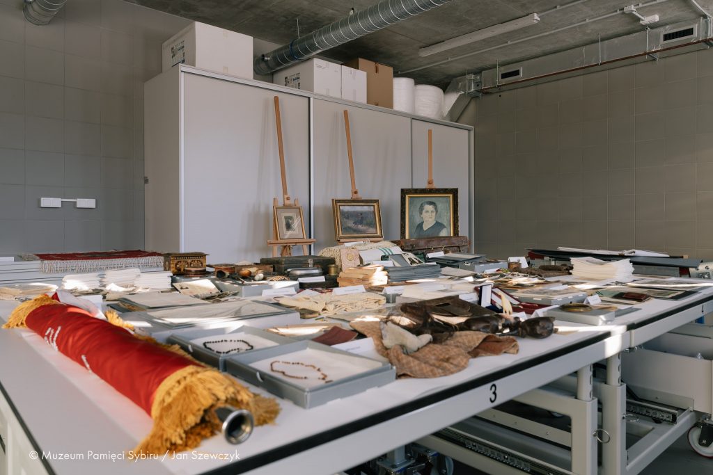 Na wielkim stole w pracowni konserwatorskiej Muzeum Pamięci Sybiru rozłożone są różnego rodzaju obiekty: sztandar, pudełka z różańcami, ubrania, książki, puzderka, dokumenty, archiwalne fotografie. Na drugim planie sztalugi z oprawionymi w ramę obrazami.