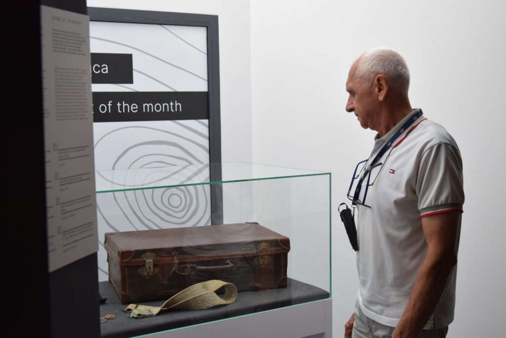 Prezentacja Eksponatu miesiąca w Muzeum Pamięci Sybiru. Z prawej strony zdjęcia mężczyzna patrzący na gablotę (z lewej) zawierającą walizkę i pas.