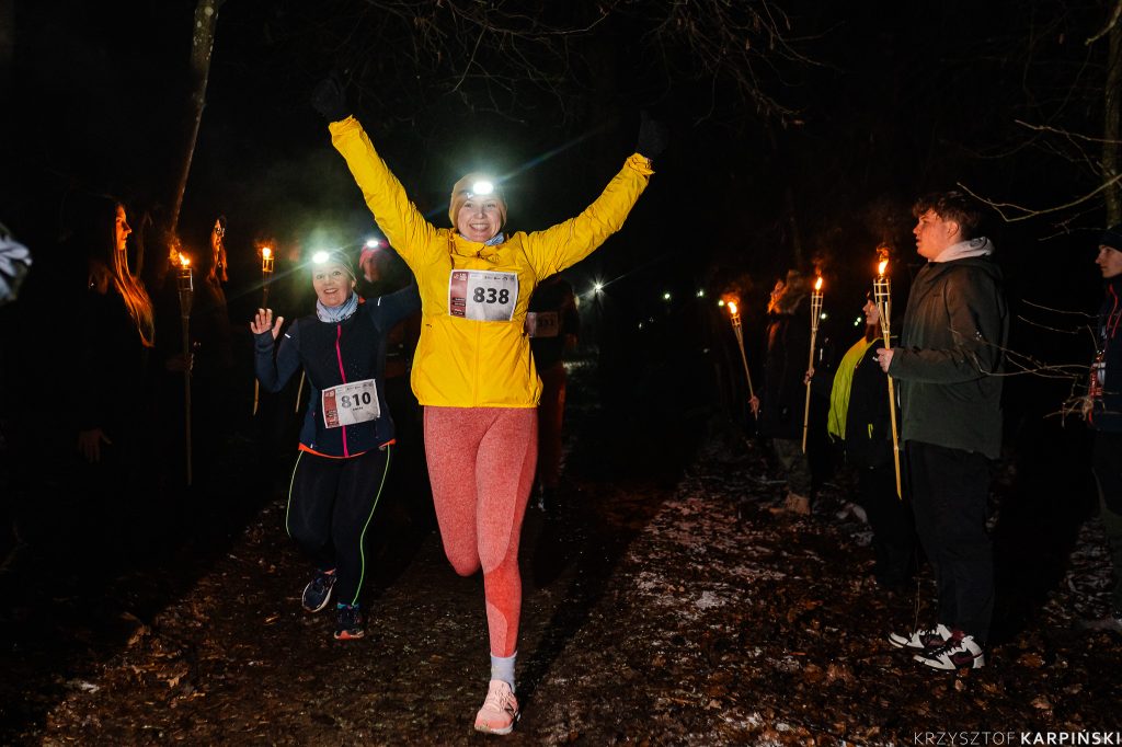 Uśmiechnięta kobieta w żółtej kurtce sportowej i różowych legginsach unosi ręce do góry podczas biegu. Za nią widać innych biegaczy. Tuż obok znajdują się osoby, które trzymają zapalone świeczki.