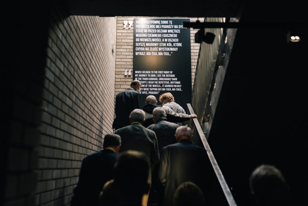 Grupa zwiedzających wchodzi po schodach na drugie piętro wystawy Muzeum Pamięci Sybiru. U góry schodów na ścianie duża czarna tablica z cytatem ze wspomnień Sybiraka.