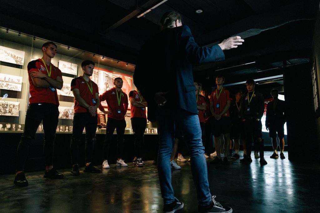 Grupa sportowców w czerwonych koszulkach oprowadzana po wystawie stałej Muzeum Pamięci Sybiru przez przewodnika