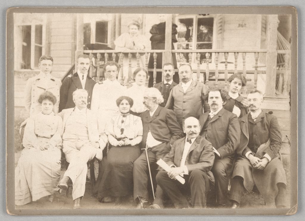 Czarno-biała fotografia z początku XX wieku, wykonana w Bołogoje, Rosja. Grupa kobiet i mężczyzn siedzi lub stoi przed tarasem drewnianego domu.