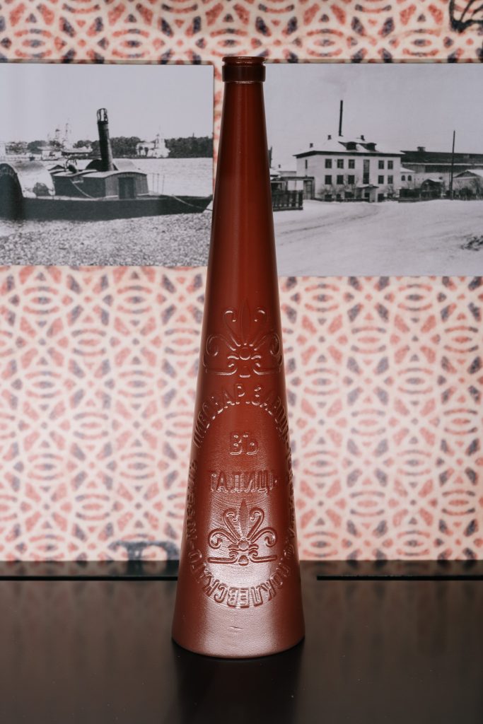 Gablota na wystawie stałej Muzeum Pamięci Sybiru. Na tle czarno-białych zdjęć i wzorzystej tapety prezentowana jest kopia wysokiej i smukłej, brązowej butelki po piwie.