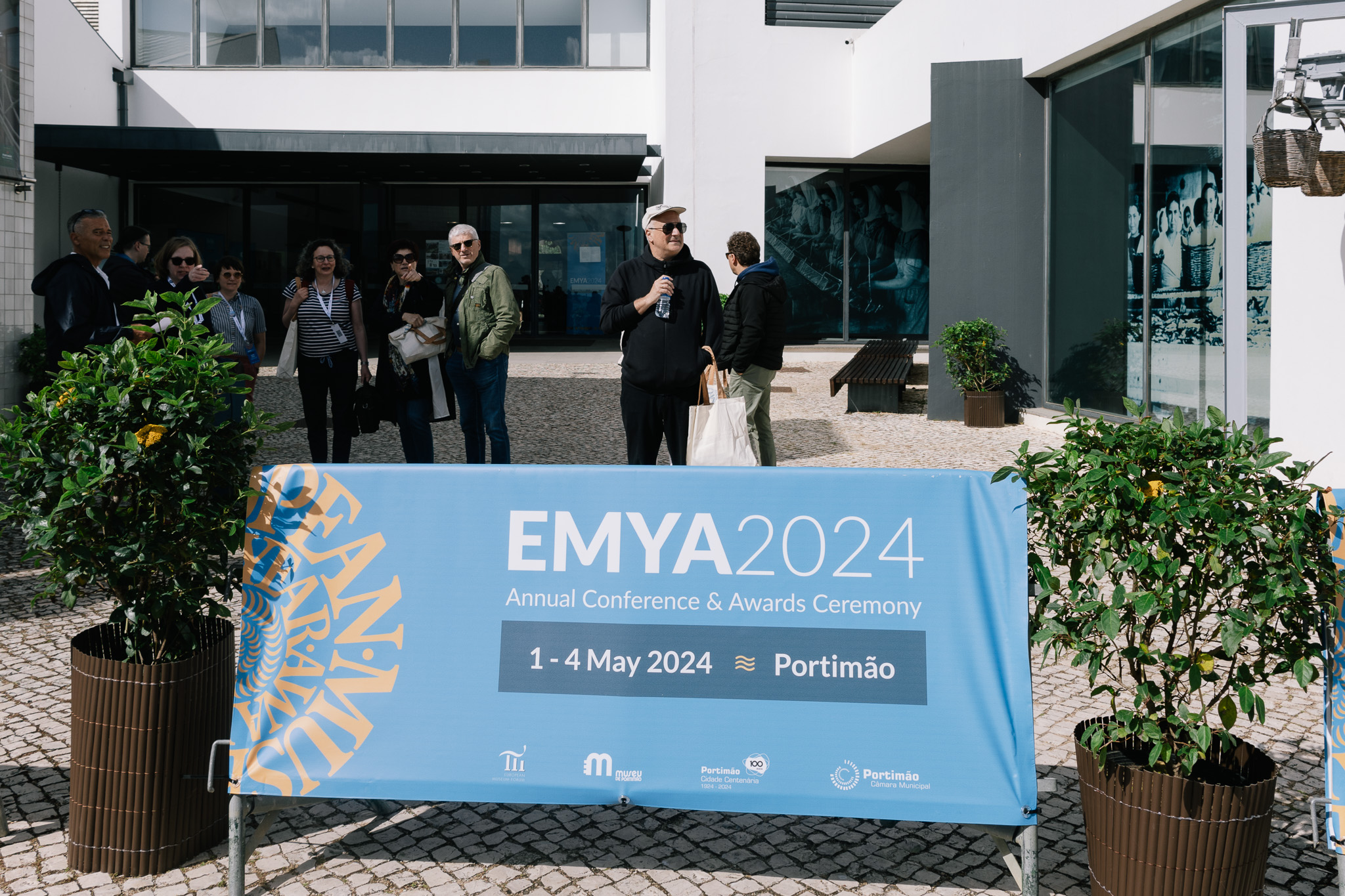 Zdjęcie przedstawia plakat gali EMYA 2024 oraz uczestników gali stojących przed budynkiem.