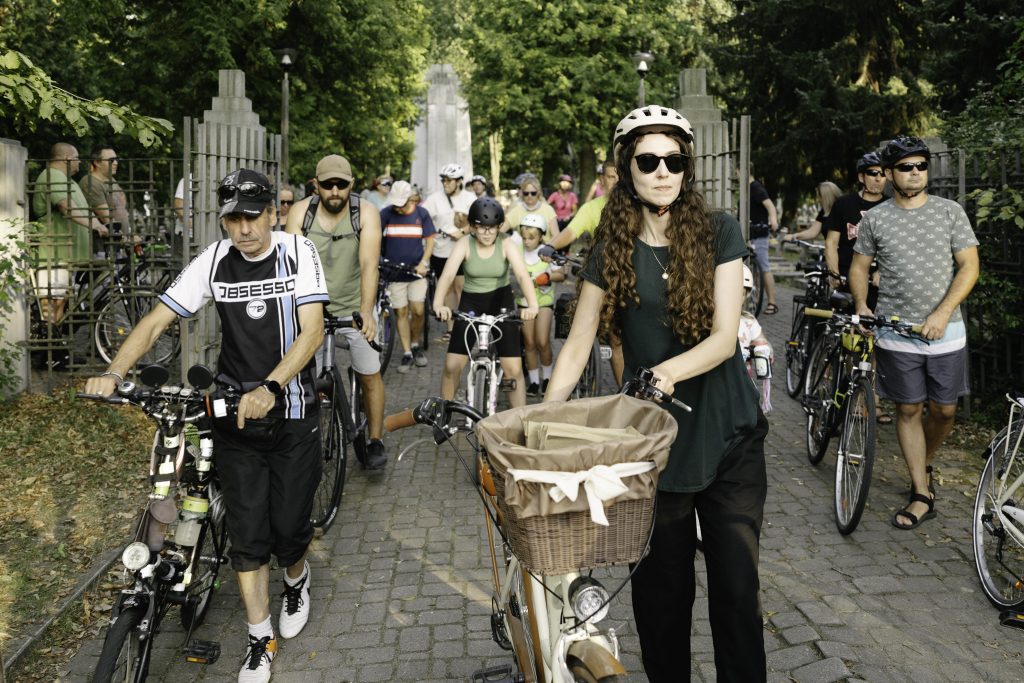 Duża grupa uczestników Peletonu wychodzi z Cmentarza Wojskowego, prowadząc swoje rowery. Na pierwszym planie młoda kobieta z długimi, kręconymi włosami.