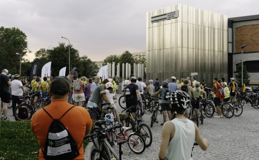 Duża grupa uczestników Peletonu stoi przy swoich rowerach na tle wejścia głównego do Muzeum Pamięci Sybiru.