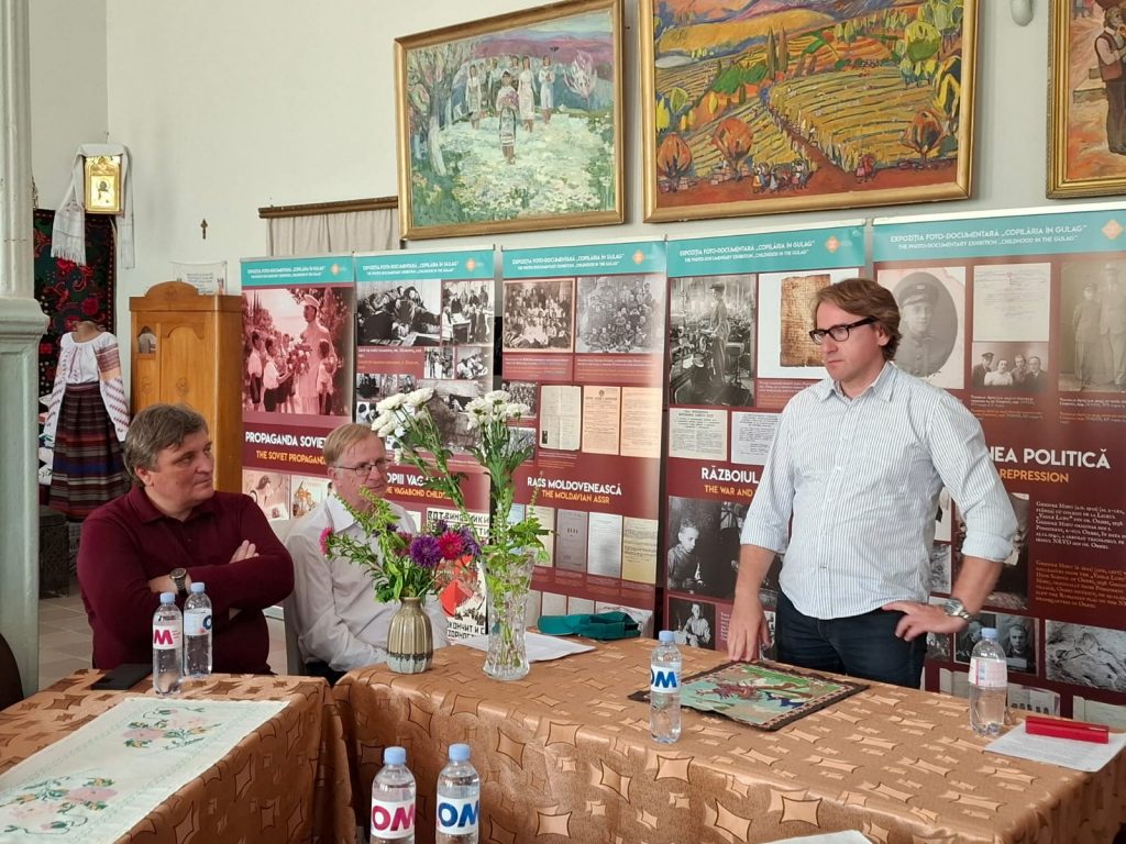 Dyrektor Muzeum Pamięci Sybiru prof. Wojciech Śleszyński przemawia do siedzących osób. Za nim znajduje się ekspozycja Muzeum Narodowego Historii Mołdawii na temat dzieciństwa mołdawskich dzieci w miejscach deportacji
