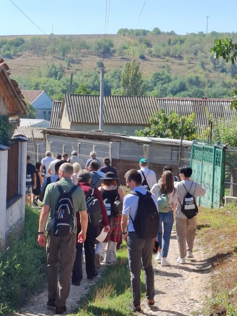 Grupa uczestników Peletonu idzie polną drogą. Obok są zabudowania.