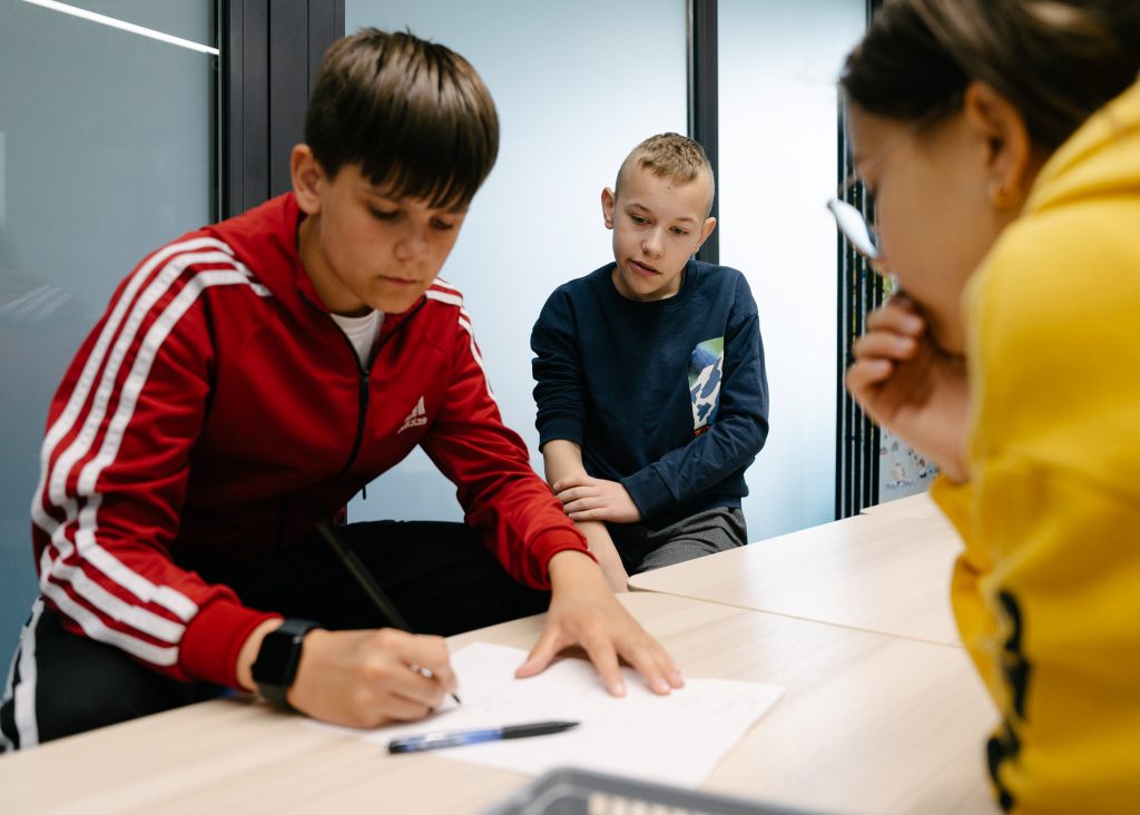 Kilkunastoletni chłopiec siedzi przy stole i skupiony notuje. Obok siedzi inny chłopiec i dziewczynka, z uwagą przyglądają się