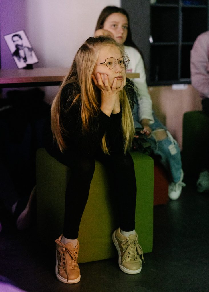 Kilkunastoletnia dziewczynka siedzi na pufie. Twarz opiera na dłoniach