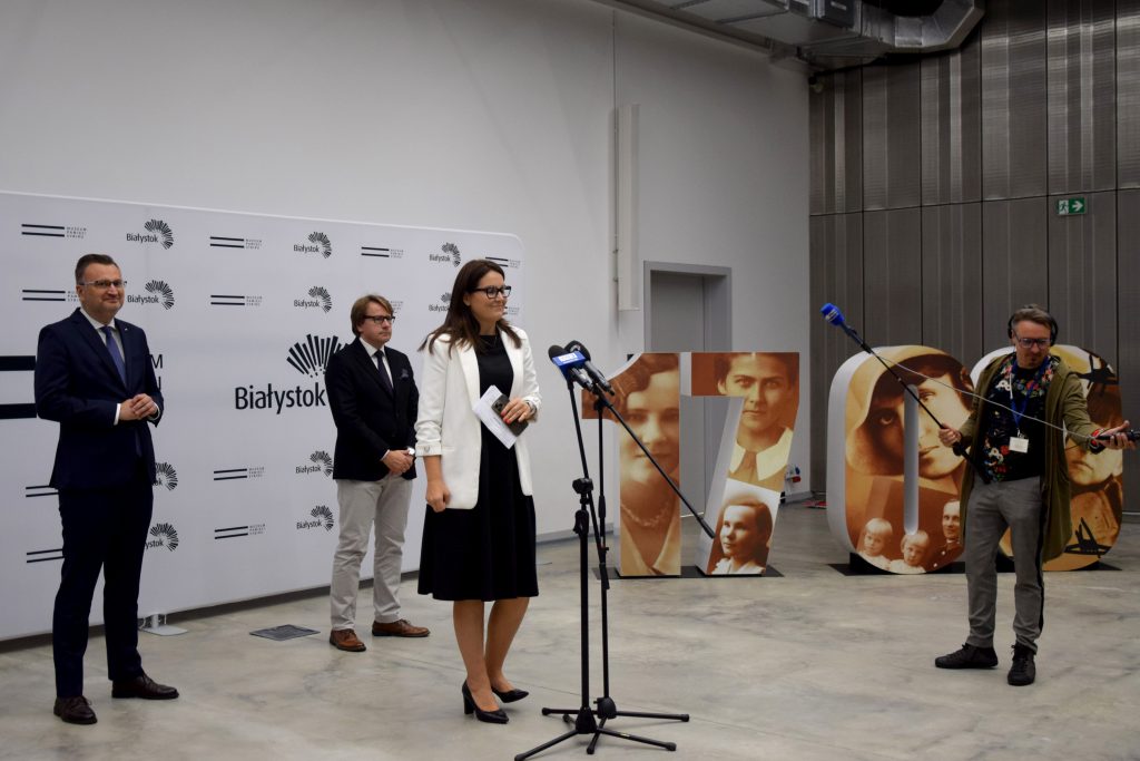 Rzecznik prasowy prezydenta Białegostoku przemawia do mikrofonu podczas konferencji prasowej. Z tyłu stoją zastępca prezydenta miasta Rafał Rudnicki oraz Dyrektor Muzeum Pamięci Sybiru prof. Wojciech Śleszyński.  