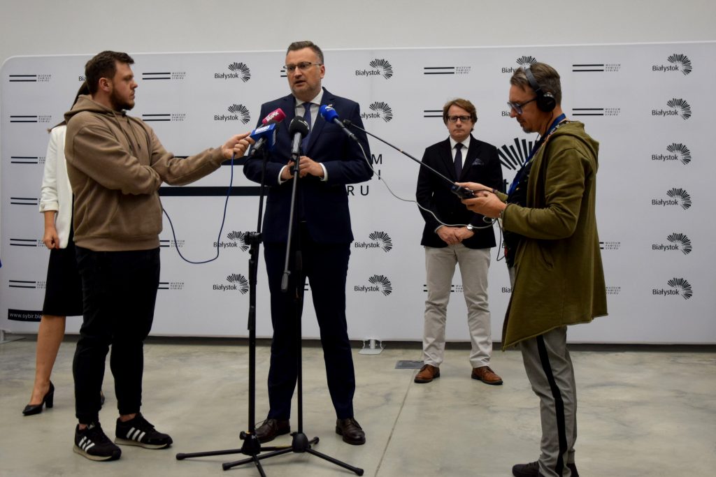 Dyrektor Muzeum Pamięci Sybiru prof. Wojciech Śleszyński przemawia do mikrofonu podczas konferencji prasowej. Obok stoi dziennikarz oraz zastępca prezydenta miasta Rafał Rudnicki i rzecznik prasowy prezydenta Tadeusza Truskolaskiego.