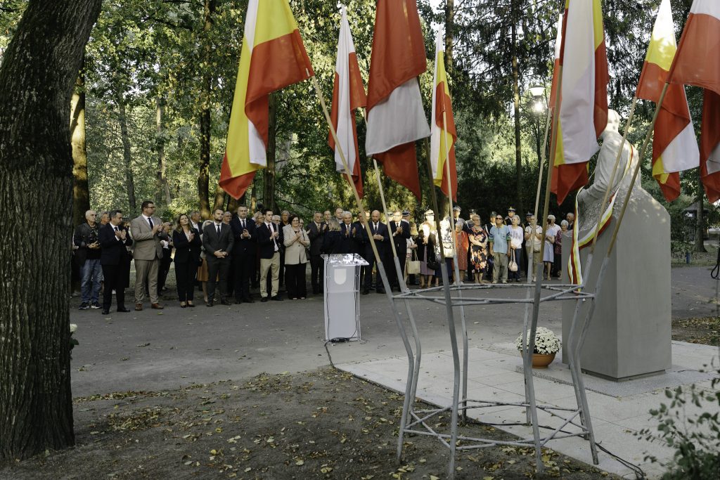 Na pierwszym planie flagi Polski i Białegostoku, za flagami pomnik Seweryna Nowakowskiego. Z tyłu grupa uczestniczących w uroczystości.
