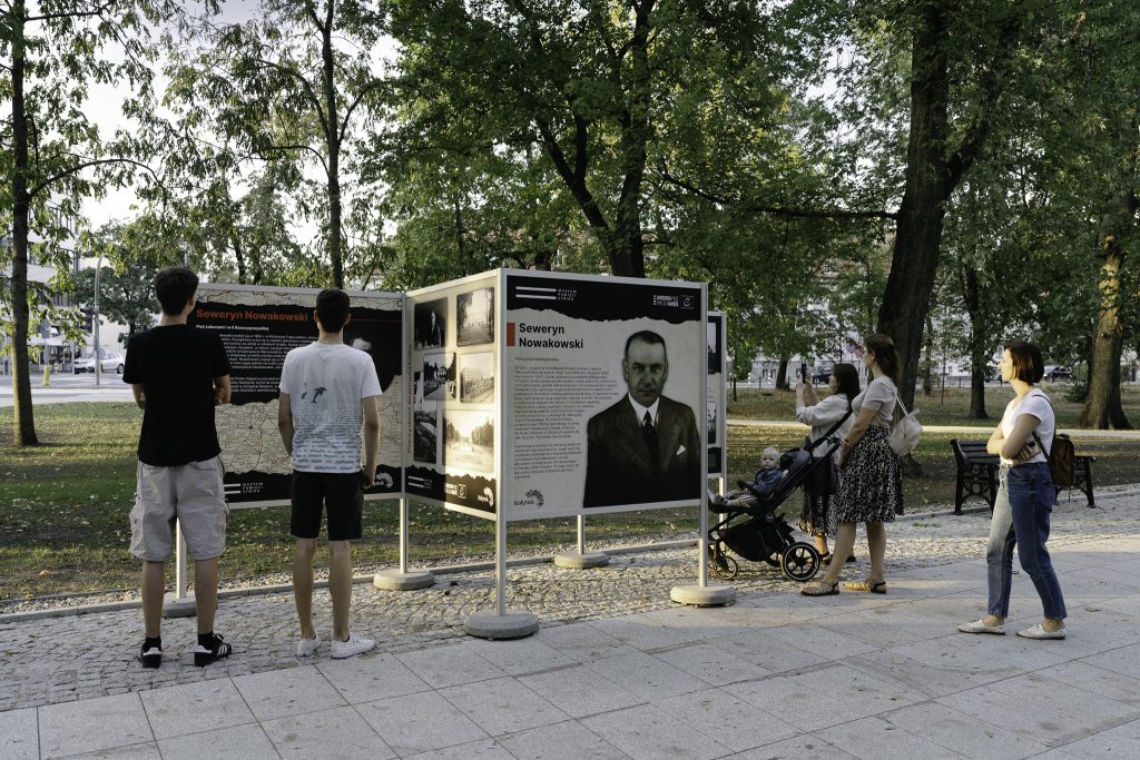 Grupa osób przygląda się wystawie plenerowej poświęconej Sewerynowi Nowakowskiemu.