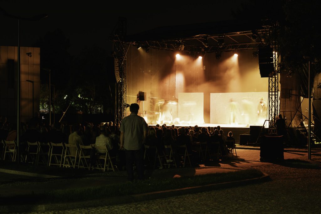 Oświetlona scena podczas spektaklu. Przed sceną widzowie siedzący na krzesłach.