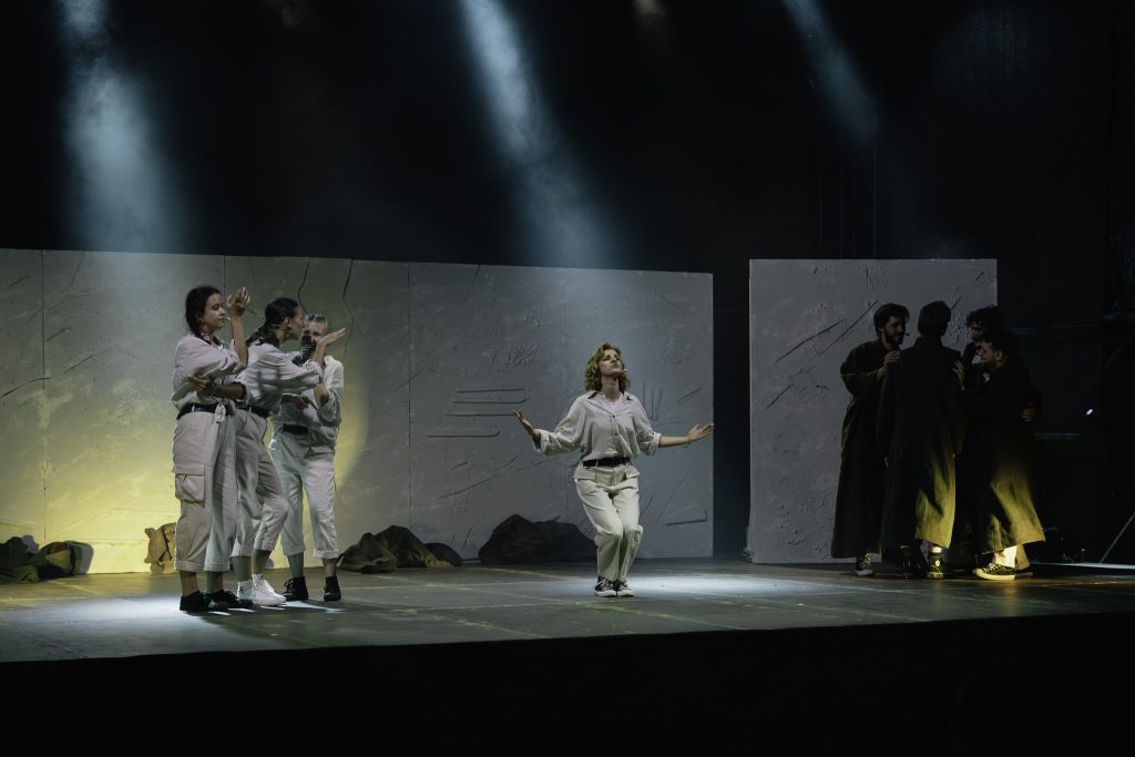 Aktorka śpiewa na scenie. Ma ugięte nogi i rozłożone ręce. Z lewej strony grupa dziewcząt, z prawej strony grupa młodych mężczyzn.