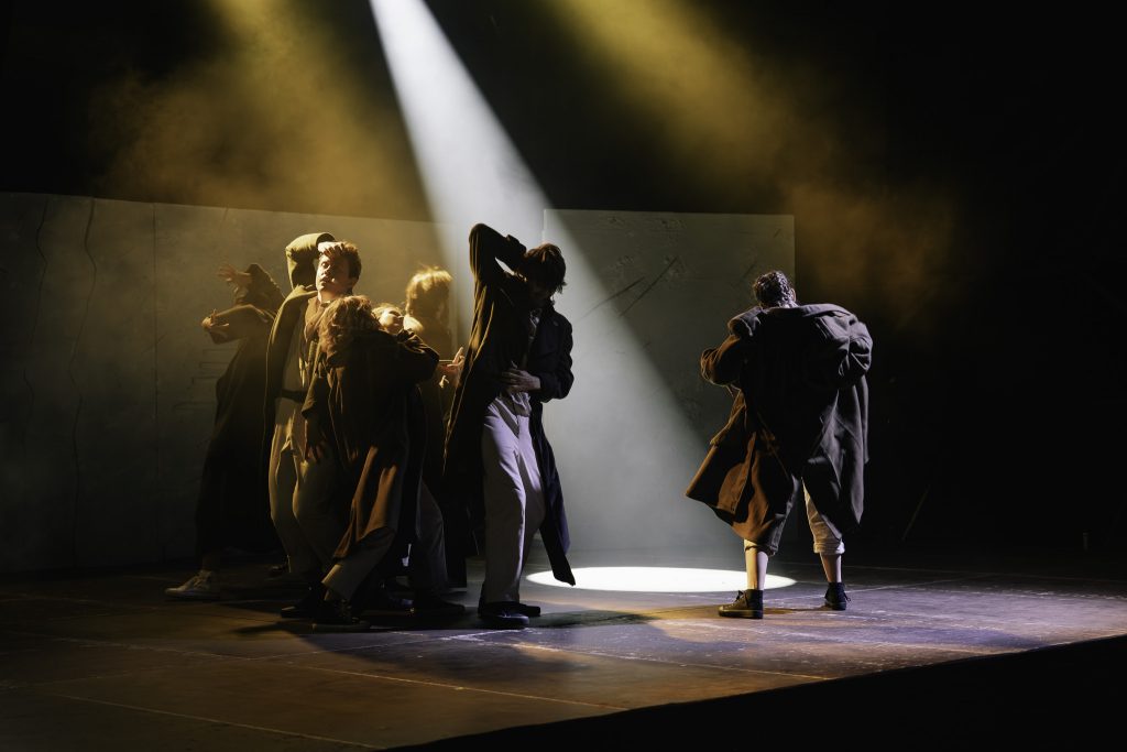 Aktorzy na scenie podczas spektaklu. Ubrani są w długie płaszcze i jasne spodnie.