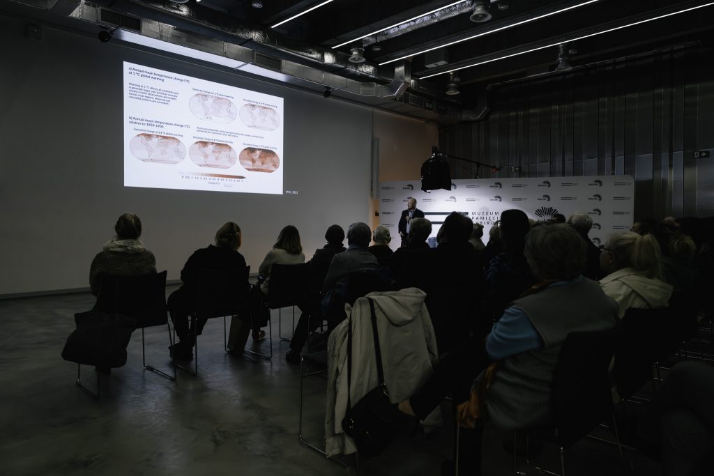 Grupa uczestników spotkania słucha wykładu. Widoczny jest wyświetlany slajd oraz stojący w oddali prof. Mariusz Lamentowicz.