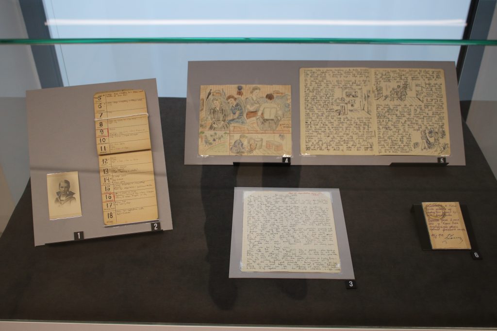 Eksponaty w szklanej gablocie: portretowe zdjęcie nastoletniej dziewczynki, dziennik z wpisami, kolorowy rysunek, dokument oraz odręczne zapiski.