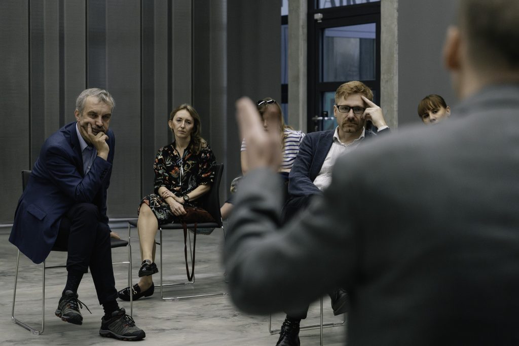 Grupa osób siedzi i słucha przemawiającego naprzeciw nich mężczyzny.