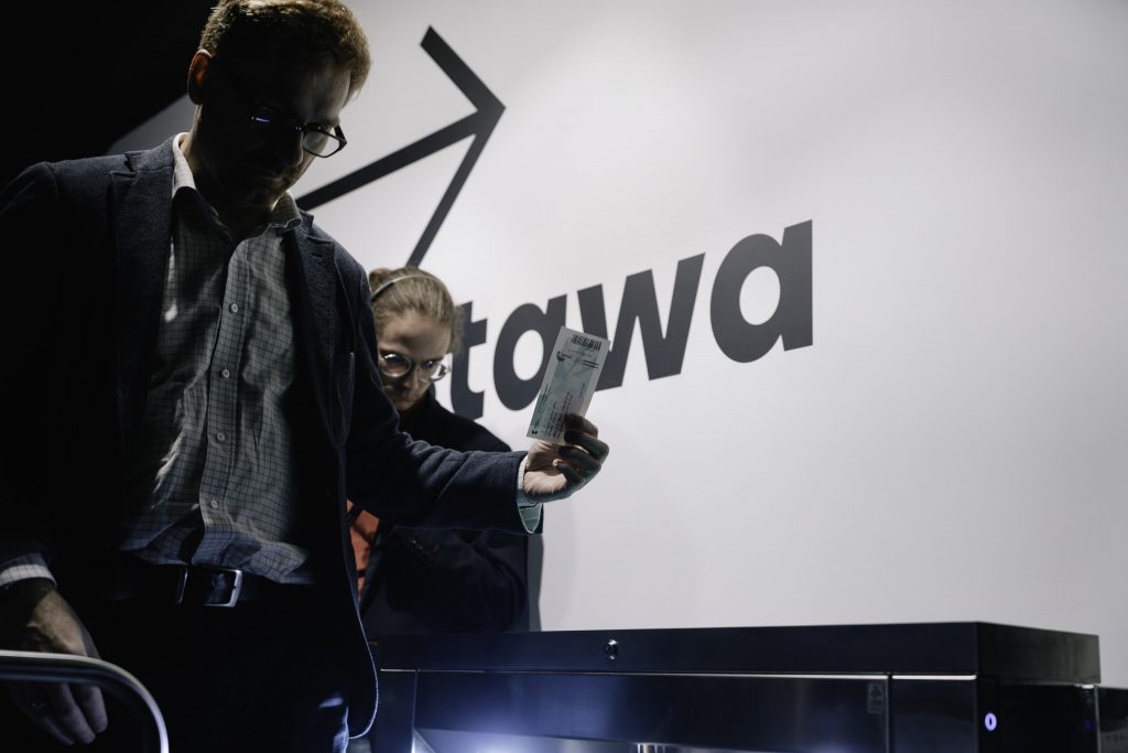 Mężczyzna przechodzi przez bramkę, w ręku trzyma bilet. Nad nim znajduje się napis "wystawa".