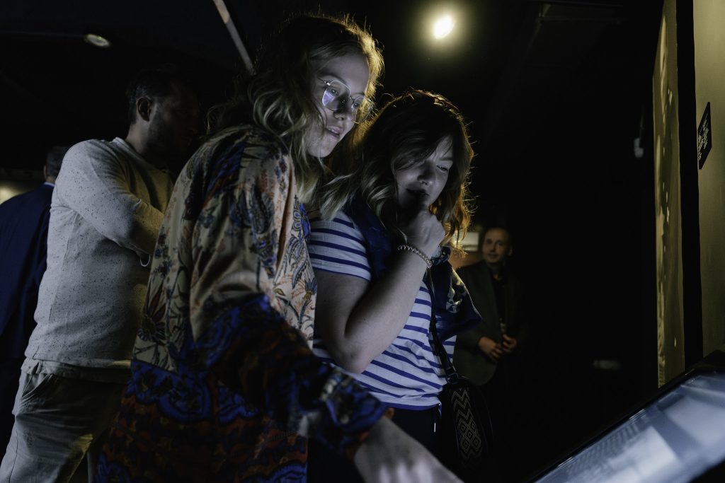 Dwie młode kobiety stoją blisko siebie i patrzą na ekran znajdujący się przed nimi. Kobieta po lewej nosi kolorową, wzorzystą kurtkę, a kobieta po prawej ma na sobie pasiastą koszulkę i trzyma rękę przy twarzy, jakby zastanawiając się nad czymś.