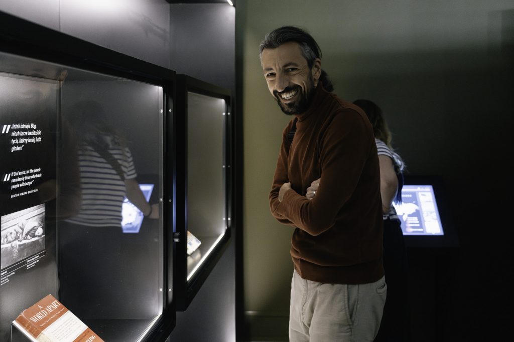 Mężczyzna w brązowym swetrze uśmiecha się i stoi z założonymi rękami. W tle są gabloty z eksponatami i tekstem, a także ekran dotykowy,