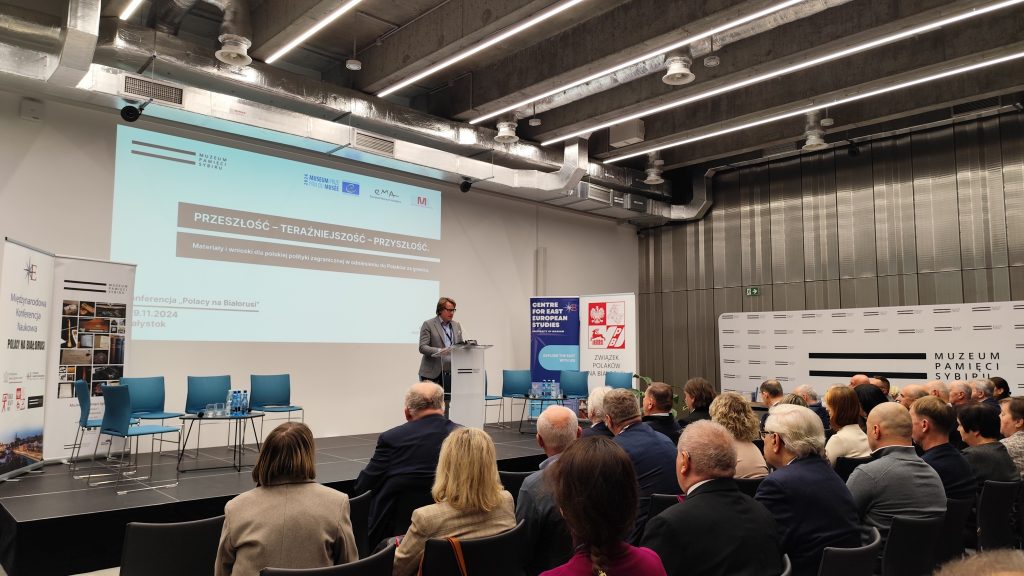 Prof. Wojciech Śleszyński przemawia do zgromadzonych uczestników konferencji. Dyrektor stoi przy mównicy, za nim w tle wyświetlany jest slajd z informacją o konferencji. Uczestnicy konferencji siedzą na krzesłach i uważnie słuchają.