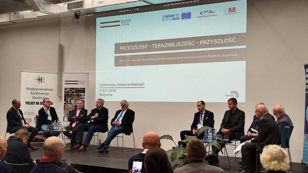 Na scenie siedzi 10 osób. Jedna z nich mówi do mikrofonu. Przy nich stoją dwa stoliki na których znajdują się szklanki i butelki z wodą. W tle na ścianie wyświetlany jest slajd z informacją o konferencji.