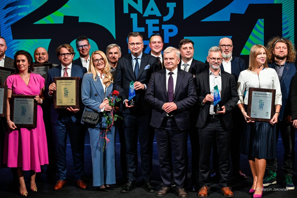 Grupa osób stoi na scenie. Niektóre z nich trzymają w rękach certyfikaty lub statuetki. Wśród nich prof. Wojciech Śleszyński trzyma certyfikat "Najlepszy", obok niego Rafał Rudnicki trzyma statuetkę "Najlepszy".