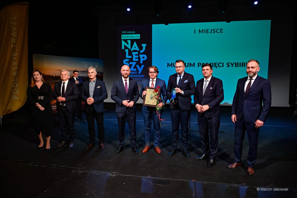 Grupa osób stoi na scenie. Wśród nich, prof. Wojciech Śleszyński, trzyma certyfikat "Najlepszy" oraz różę, obok niego Rafał Rudnicki trzyma statuetkę "Najlepszy".