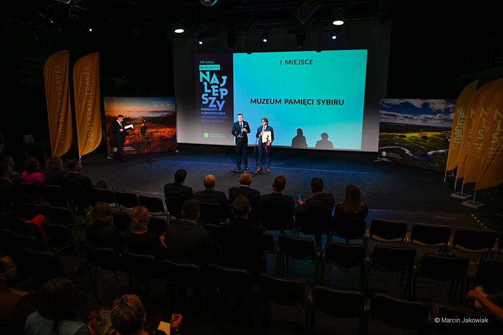 Na scenie przy mikrofonach stoją dyrektor Muzeum Pamięci Sybiru, prof. Wojciech Śleszyński oraz zastępca prezydenta Białegostoku, Rafał Rudnicki. Prof. Wojciech Śleszyński  w rękach trzyma certyfikat "Najlepszy" oraz różę, obok niego Rafał Rudnicki trzyma statuetkę "Najlepszy". W tle na dużym ekranie wyświetlany jest slajd "1 miejsce Muzeum Pamięci Sybiru".