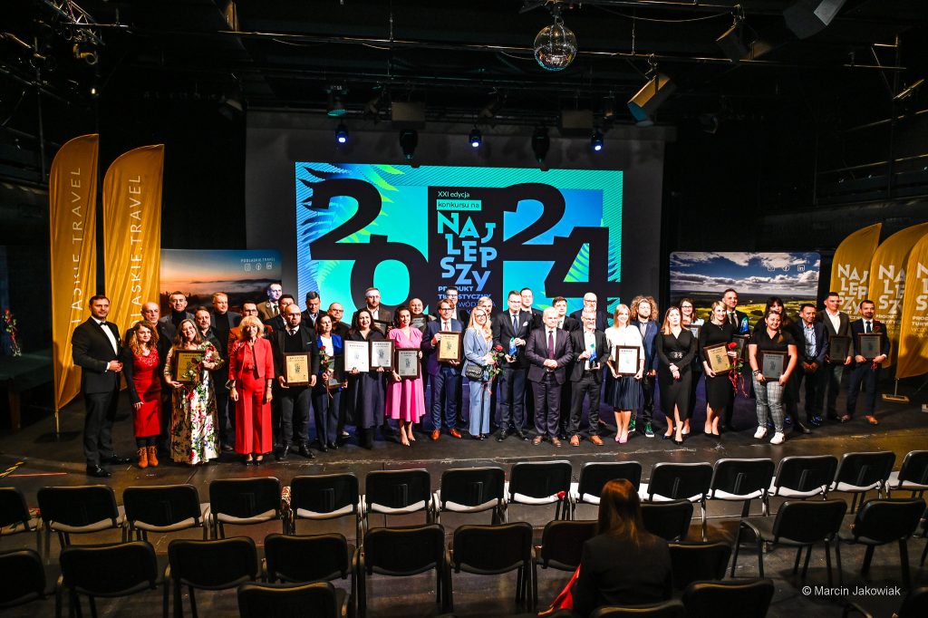 Na scenie stoi grupa kilkudziesięciu osób. Wiele z nich trzyma w rękach certyfikaty. Dwie z nich trzymają w rękach statuetki. W tle na ekranie widoczny jest slajd konkursu na najlepszy produkt turystyczny województwa podlaskiego.