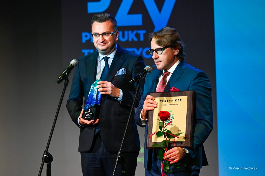 Dwie osoby stoją przy mikrofonach. Prof. Wojciech Śleszyński przemawia do mikrofonu, w rękach trzyma certyfikat "Najlepszy" oraz różę, obok niego Rafał Rudnicki trzyma statuetkę "Najlepszy".