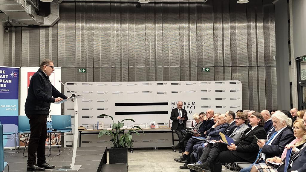 Jarosław Książek – konsul generalny RP w Grodnie przemawia przy mównicy. Widać kilkadziesiąt osób które siedzą i słuchają przemówienia, W tle, w głębi widać stoły na których leży wiele książek. 