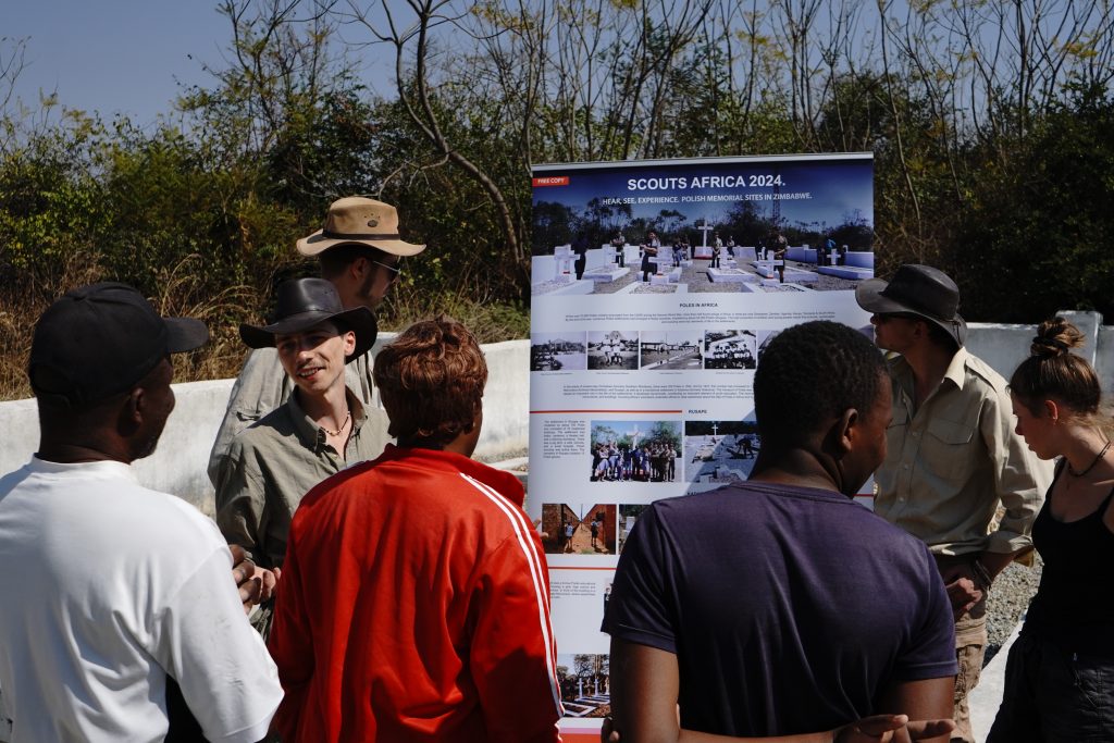 Grupa harcerzy zwraca się do zebranych wokół plakatu osób.