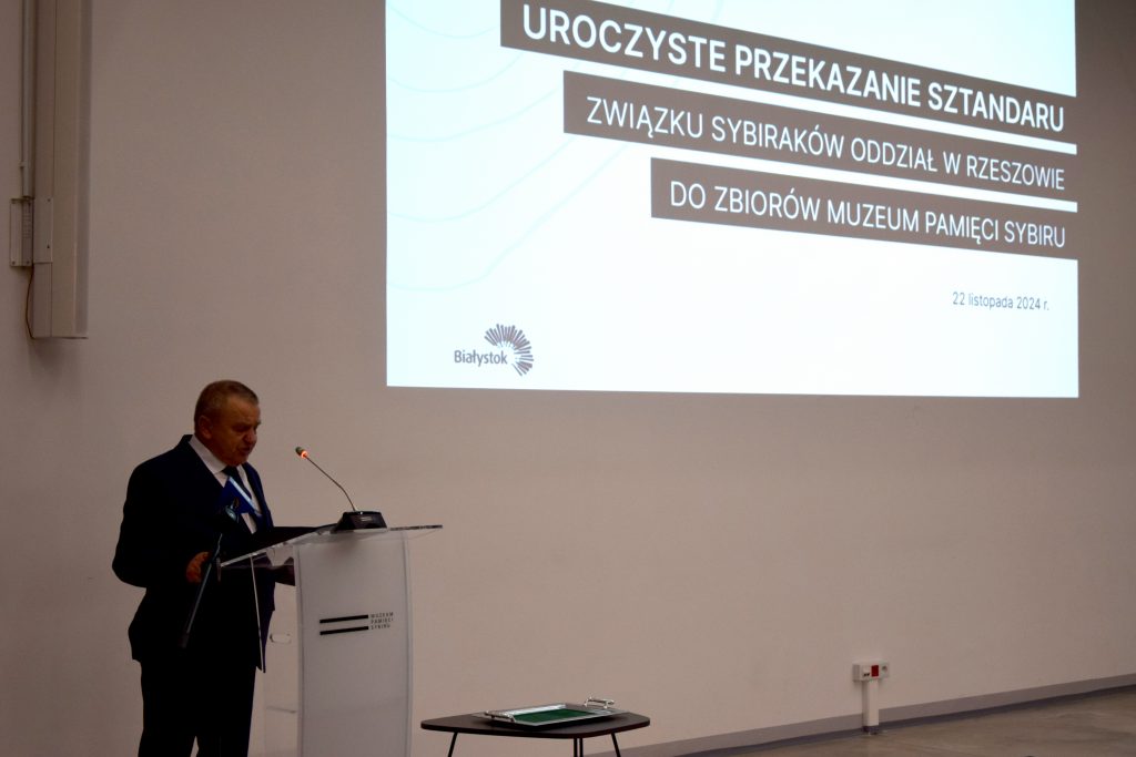 Józef Sieńko, prezes zarządu rzeszowskiego oddziału Związku Sybiraków, przemawia przy mównicy. W tle na ścianie jest wyświetlany slajd z informacją o uroczystości.