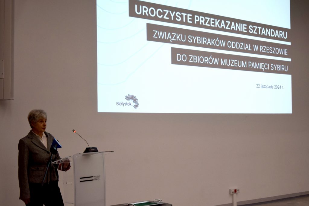 Danuta Zedania, członek zarządu rzeszowskiego oddziału Związku Sybiraków przemawia przy mównicy. W tle na ścianie jest wyświetlany slajd z informacją o uroczystości.