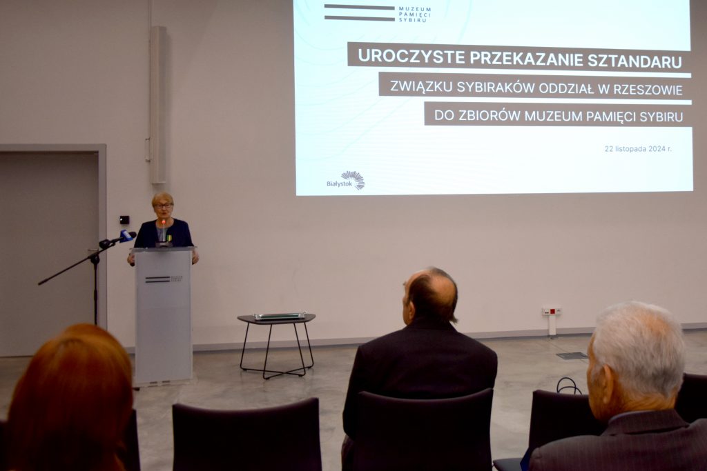 Jolanta Hryniewicka, prezes białostockiego Związku Sybiraków przemawia przy mównicy. W tle na ścianie jest wyświetlany slajd z informacją o uroczystości.