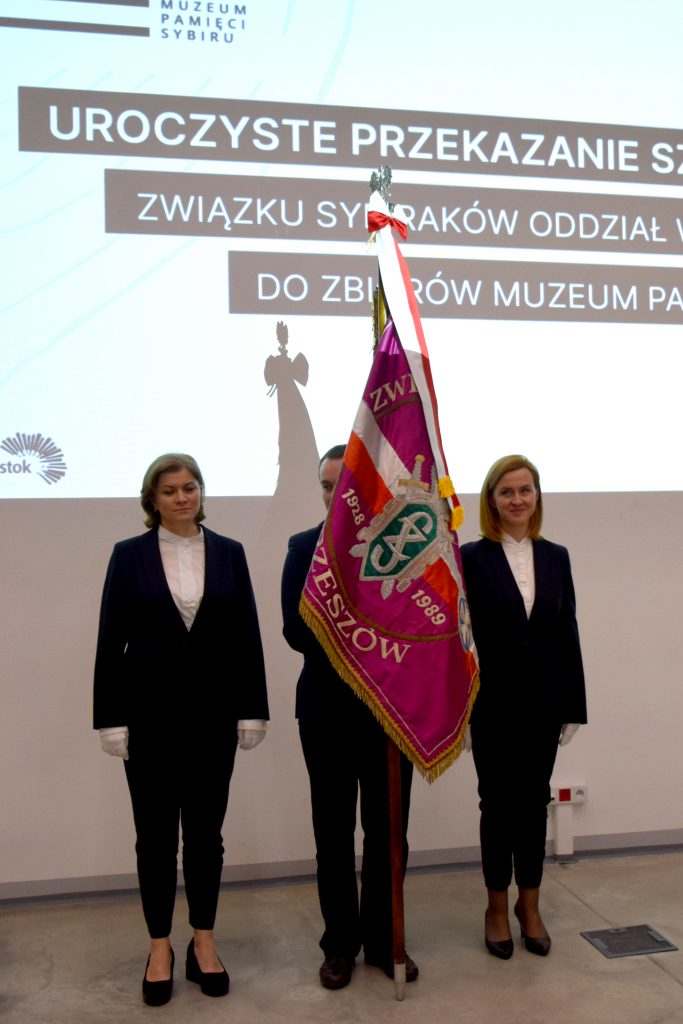 Osoby są ubrane w galowe stroje. Dwie kobiety stoją po obu stronach mężczyzny. Mężczyzna trzyma sztandar. W tle na ścianie wyświetlany jest obraz z rzutnika z informacją o wydarzeniu.